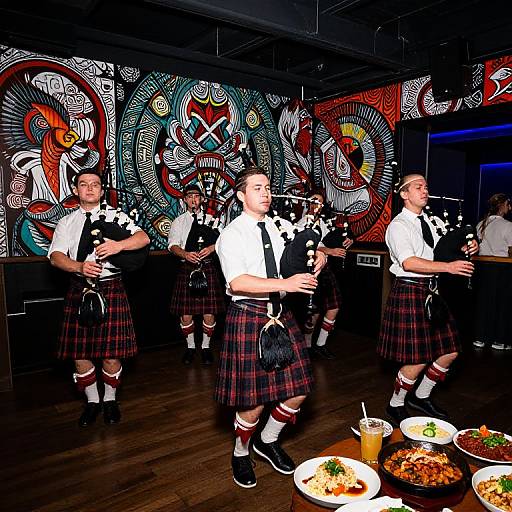 Photograph of six men in white shirts, black vests, and red tartan kilts, playing bagpipes in a dimly lit room with vibrant