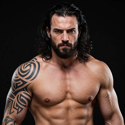 Photograph of a muscular, shirtless man with long black hair, beard, intense expression, tribal tattoo on right shoulder, against a black background.
