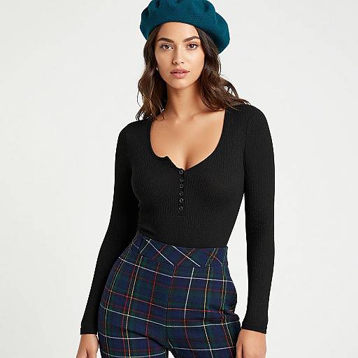 Photograph of a woman with dark wavy hair, wearing a teal beret, black ribbed long-sleeve top, and high-wa