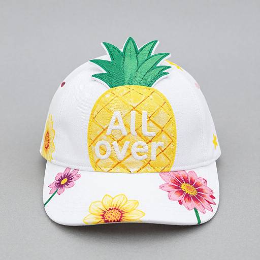 Photograph of a white baseball cap adorned with a large yellow pineapple graphic, green leaves, and pink flowers, with 