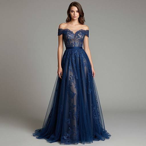 Photograph of a woman in an off-shoulder, floor-length, navy blue lace gown with floral patterns, standing against a plain gray background.