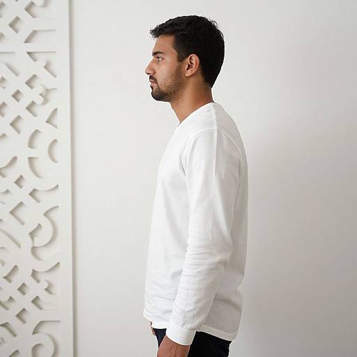 Photograph of a side-profile, dark-haired man in a white shirt, standing against a white wall with a geometric pattern on the left.
