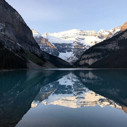 Photograph of a serene mountain lake with a clear, reflective surface mirroring snow-capped peaks and surrounding dark, rocky cliffs.