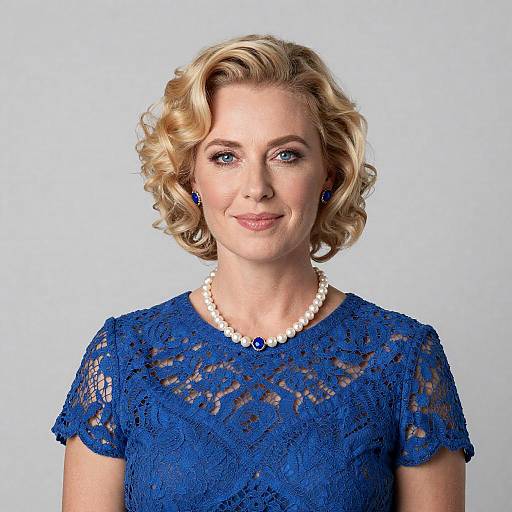Photograph of a blonde woman with curled hair, wearing a blue lace dress, white pearl necklace, and blue earrings, smiling against a plain white background