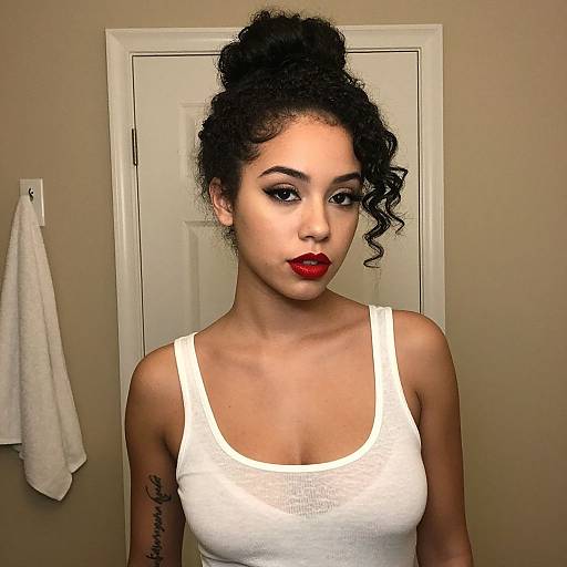 Photograph of a young woman with curly black hair in a bun, wearing a white tank top, red lipstick, and visible tattoo on her left arm