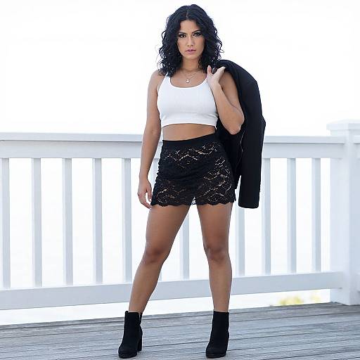 Photograph of a confident woman with curly black hair, wearing a white crop top, black lace skirt, and black ankle boots, standing on a wooden