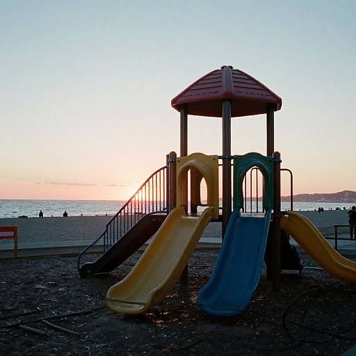 Photograph of a sunset at a beach with a colorful playground in the foreground, featuring yellow and blue slides and a red-roofed structure. Sil