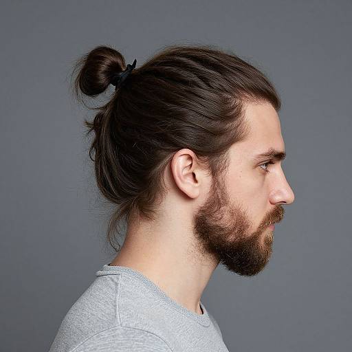 Side profile photograph of a bearded man with brown hair in a top knot, wearing a light gray shirt, against a gray background.