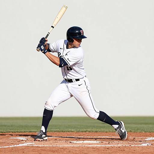 Photograph of a male baseball player in white uniform and black helmet, mid-swing, standing on a dirt field. Bright sunlight.