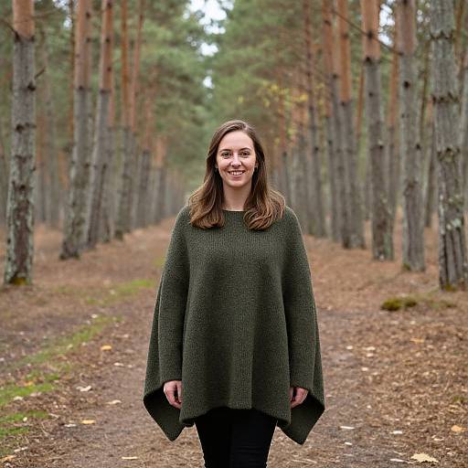 Photograph of a smiling woman with long brown hair, wearing a dark green, loose-knit sweater, standing on a forest path lined with tall pine