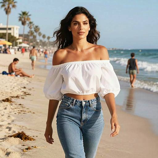 Photograph of a beautiful woman with long dark hair, off-shoulder white blouse, and high-waisted blue jeans walking on a sunny beach