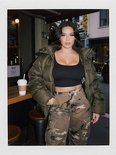 Photograph of a curvy woman with long black hair, wearing a black crop top, green puffer jacket, and camouflage pants, standing in a