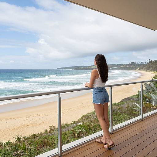 Photograph of a woman with long black hair, wearing a white tank top and blue denim shorts, standing on a wooden balcony, gazing at a