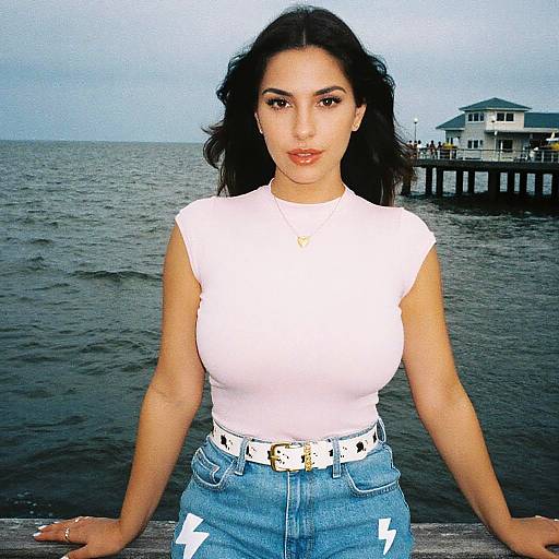 Photograph of a young woman with long dark hair, wearing a white sleeveless top, blue high-waisted jeans, white belt, standing by