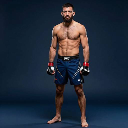 Photograph of a muscular, bearded man with short dark hair, wearing navy UFC shorts, red and white gloves, standing barefoot against a dark