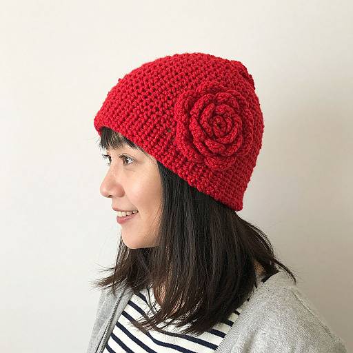 Photograph of a smiling woman with fair skin, black hair, wearing a red, crocheted knit hat with a large rose design, black-and