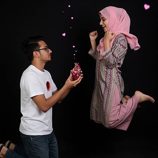 Photograph of a smiling woman in pink hijab and traditional dress, floating mid-air, as a man in white shirt and glasses offers a red apple