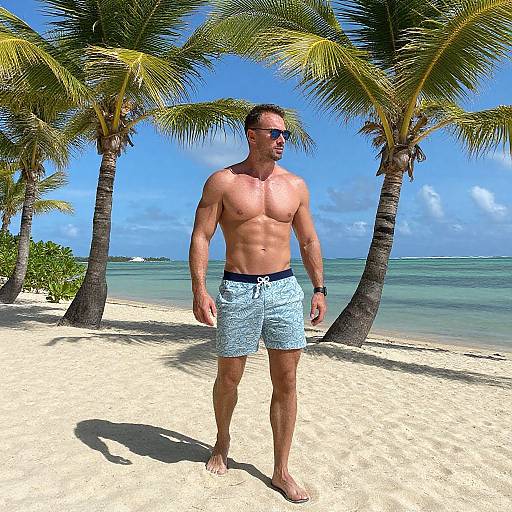 Photograph of a muscular, shirtless man in blue patterned swim trunks and sunglasses walking on a sunny tropical beach with palm trees and clear blue