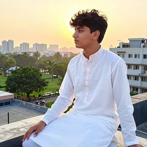 Photograph of a young man with dark curly hair, wearing a white traditional long kurta, sitting on a rooftop at sunset, cityscape and green