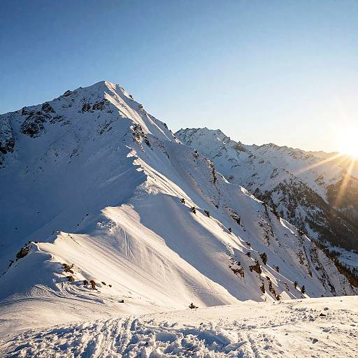 Photograph of a snow-covered mountain peak at sunrise, with bright sunlight casting long shadows, clear blue sky, and crisp, textured snow.