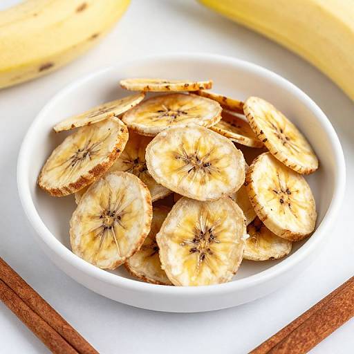 Photograph of a white bowl filled with golden-brown, sliced banana chips with visible seeds, placed on a white surface with cinnamon sticks and a banana