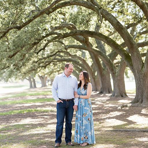 Photograph of a smiling couple standing under large, arching trees in a sunlit park. Man in checkered shirt, dark jeans; woman in