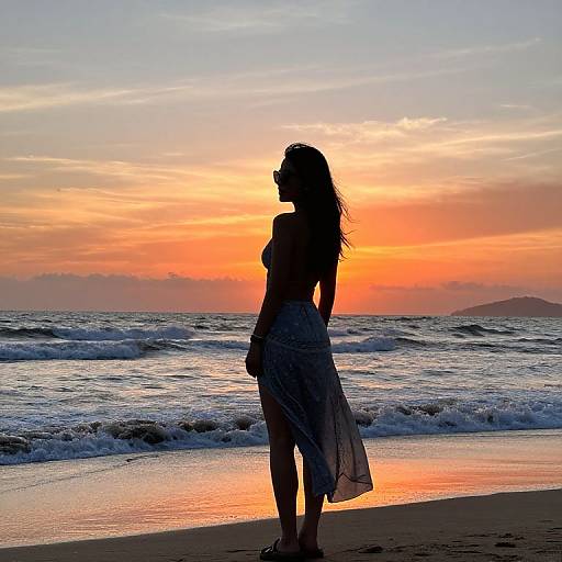 Silhouetted woman in a flowing skirt and sunglasses stands on a beach at sunset, waves gently crashing in the background.