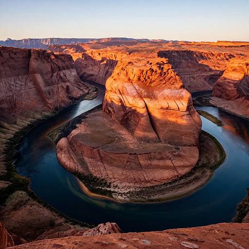 Photograph of a striking, sunlit red rock formation surrounded by a dark, winding river in a rugged canyon landscape at sunset.