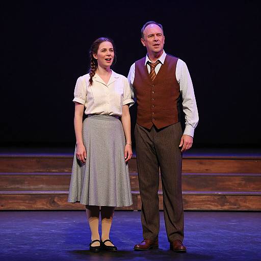 Photograph of a stage play featuring a young woman with braided brown hair, white blouse, and gray skirt, standing beside an older man in a