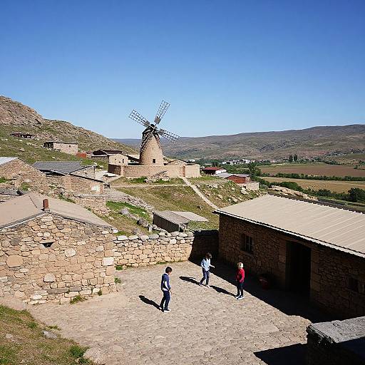Photograph of a sunny rural village with stone buildings, a windmill, and four people walking on a paved path, set against a clear blue sky
