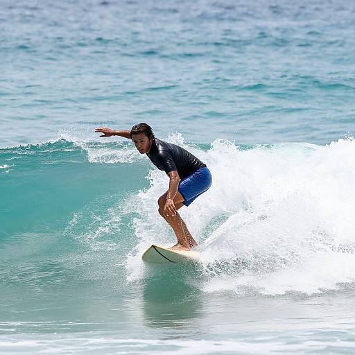Photograph of a male surfer in a black shirt and blue shorts, riding a turquoise wave with splashing white water.