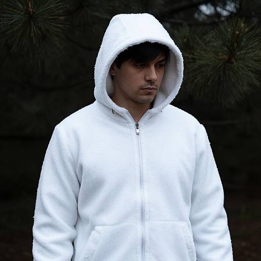 Photograph of a serious young man with dark hair, wearing a white hooded fleece jacket, standing against a dark, pine tree background.