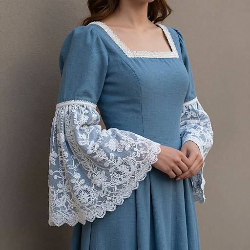 Photograph of a woman in a blue, lace-trimmed, medieval-style dress with white lace sleeves, standing against a plain gray wall.