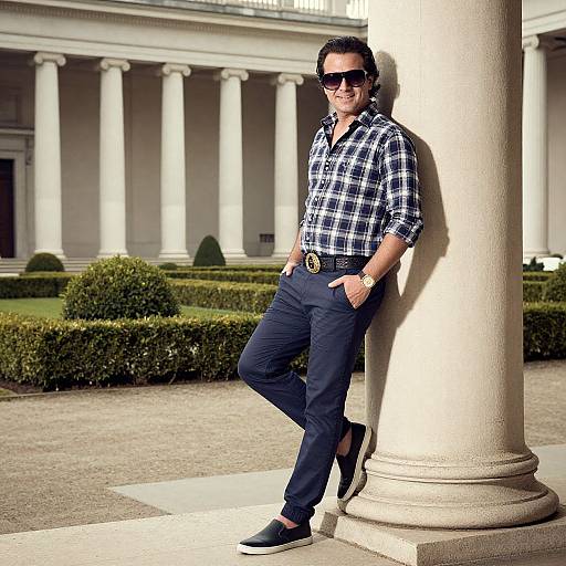 Photograph of a man in a plaid shirt, dark pants, black shoes, sunglasses, leaning against a column in a colonial-style courtyard.