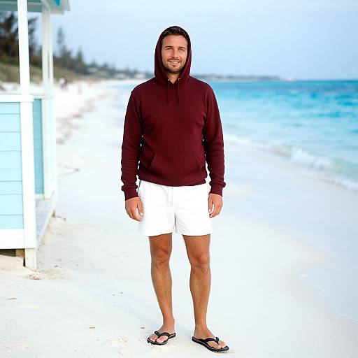 Photograph of a bearded man with long dark hair, wearing a maroon hoodie, white shorts, and black flip-flops, standing on a