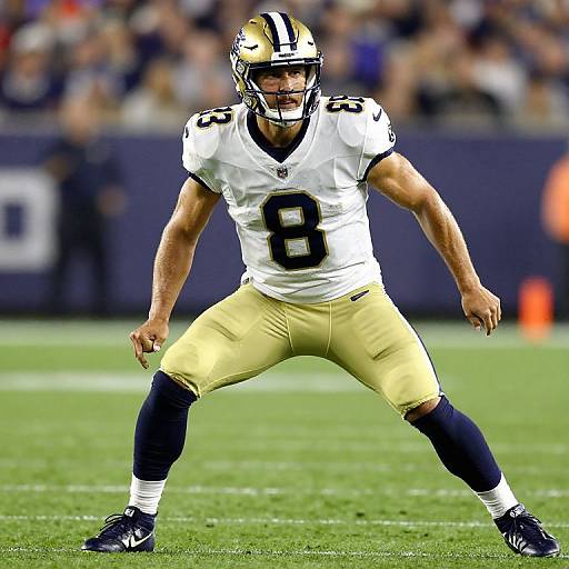 Photograph of a muscular male football player in a white and gold New Orleans Saints uniform, number 8, standing with legs apart on a green field
