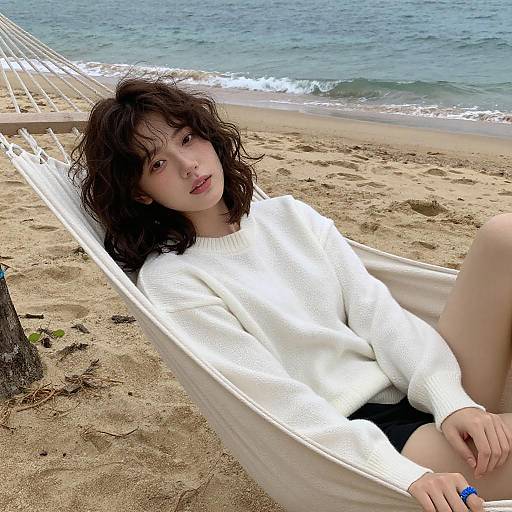 Photograph of a young woman with wavy brown hair, pale skin, and red lipstick, lounging in a white hammock on a sandy beach