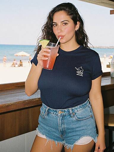 Photograph of a young woman with wavy dark hair, wearing a black t-shirt and frayed denim shorts, sipping a pink drink with a
