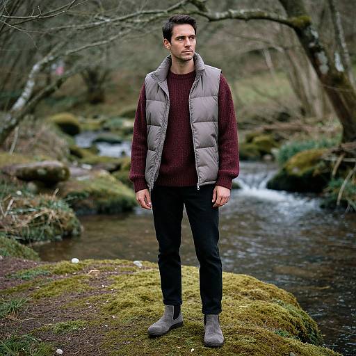 Photograph of a young man with short dark hair, wearing a gray puffer vest, maroon sweater, black pants, and gray boots, standing