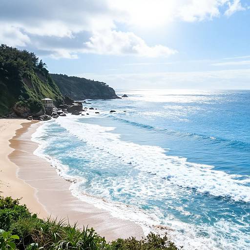 Photograph of a sunlit, curving beach with golden sand, blue waves, lush green cliffs, and a partly cloudy sky.