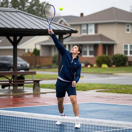 Photograph of a young male tennis player in a black athletic outfit, mid-serve on a blue tennis court, with suburban houses and a covered bench