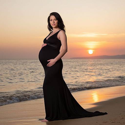 Photograph of a pregnant woman in a black, sleeveless, V-neck gown standing on a beach at sunset, with waves and a glowing orange sky