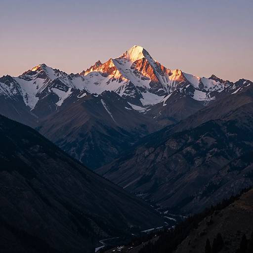 Photograph of a snow-capped mountain peak bathed in sunset light, with dark, shadowy valleys in the foreground and a clear, pastel