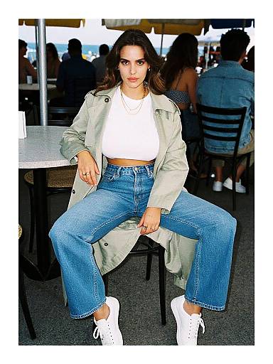 Photograph of a woman with long dark hair, light skin, wearing a white crop top, green jacket, blue high-waisted jeans, and