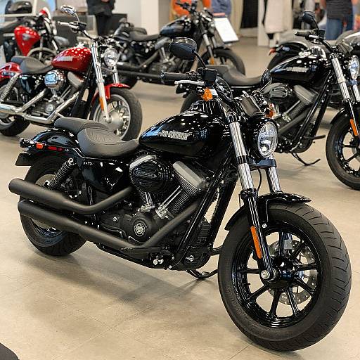 Photograph of a showroom displaying black motorcycles with shiny chrome accents, black leather seats, and thick tires, alongside a red motorcycle in the background. People