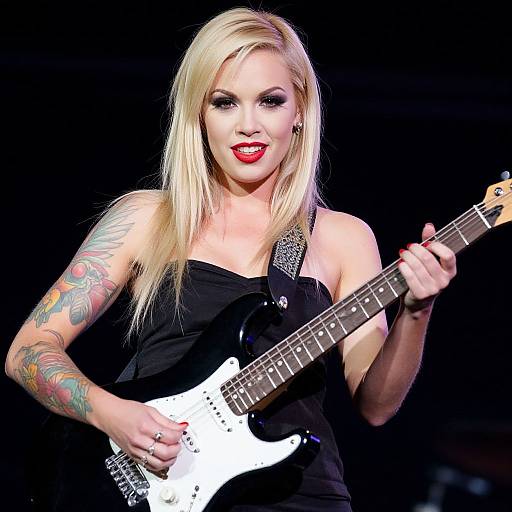 Photograph of a blonde woman with tattoos, red lipstick, black sleeveless top, playing a white electric guitar on a dark stage.