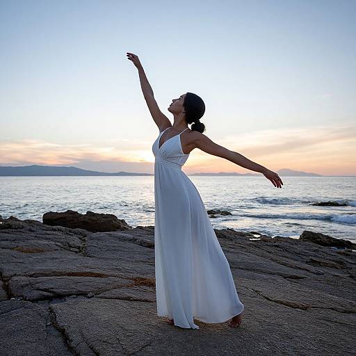 Silhouetted woman in flowing white dress dances by rocky ocean shore at sunset, arm raised, one arm extended, serene and graceful.
