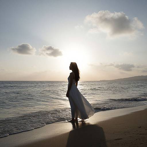 Silhouetted woman in flowing white dress walks on sunlit beach at sunset, waves gently touching shore, with clouds in the background.