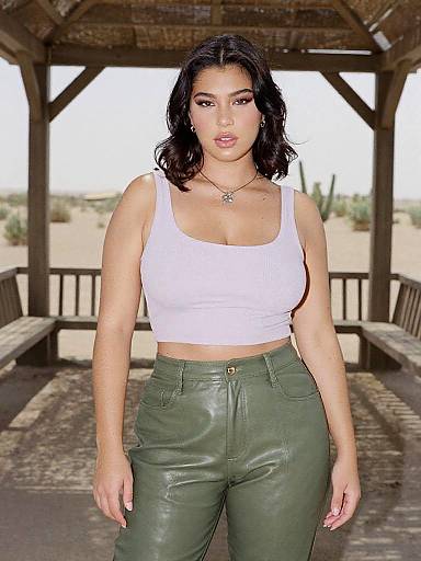 Photograph of a Latina woman with dark hair, wearing a white crop top and green leather pants, standing in a desert wooden gazebo.
