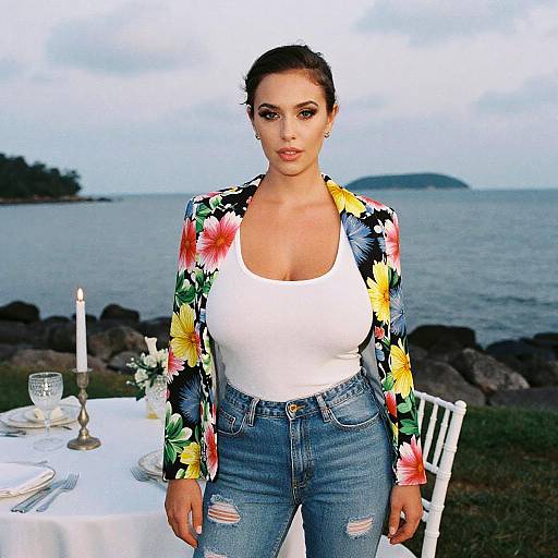 Photograph of a brunette woman with fair skin, wearing a white tank top, floral jacket, and ripped jeans, standing by a lakeside table with